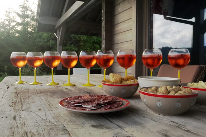 Demi-douzaine de verres d'Aperol spritz alignés sur la table de la terrasse du chalet, avec quelques apéritifs, avec vue sur les sapins et la vallée du Grand Valtin