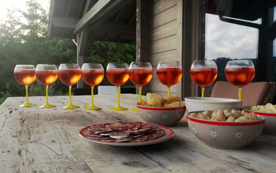 Demi-douzaine de verres d'Aperol spritz alignés sur la table de la terrasse du chalet, avec quelques apéritifs, avec vue sur les sapins et la vallée du Grand Valtin