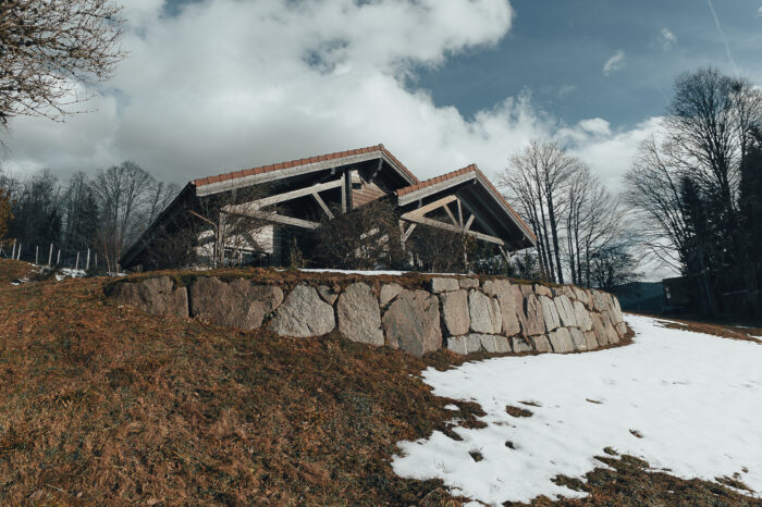 Photo 3/4 avant du chalet, prise d'en dessous de la butte, avec herbe d'hiver sechée et tâche de neiges, puis pierres de terrassement, puis le chalet posé, gris au tuiles rouges posé sur cette butte.