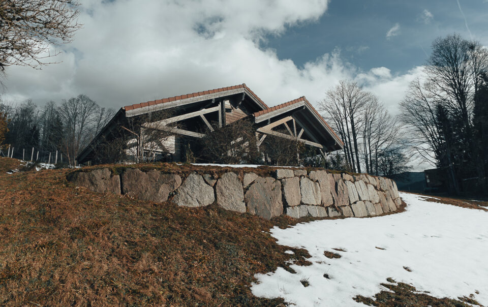 Photo 3/4 avant du chalet, prise d'en dessous de la butte, avec herbe d'hiver sechée et tâche de neiges, puis pierres de terrassement, puis le chalet posé, gris au tuiles rouges posé sur cette butte.